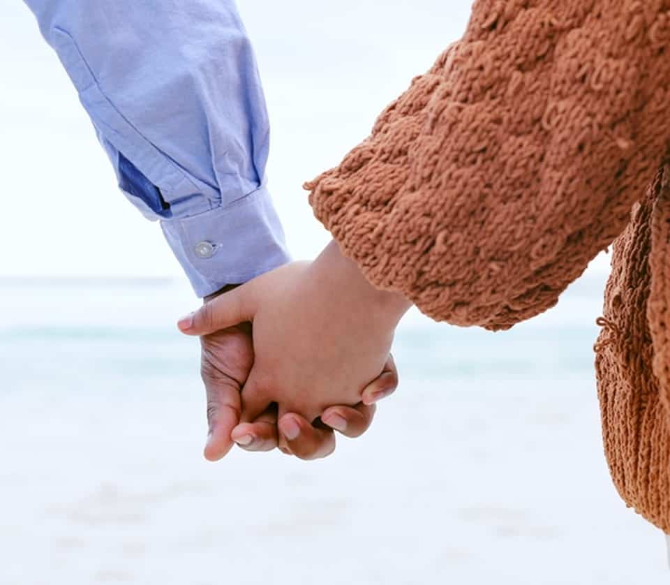 Two people holding hands on the beach, with the ocean in the background.