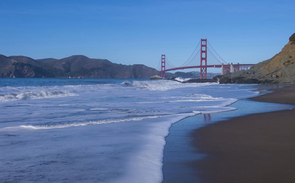 View of Golden Gate Bridge with ocean waves and sandy beach in foreground.