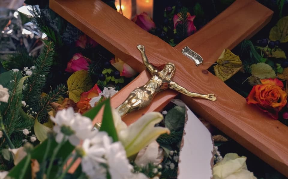 Close-up of a gold crucifix on a wooden cross with flowers around it.
