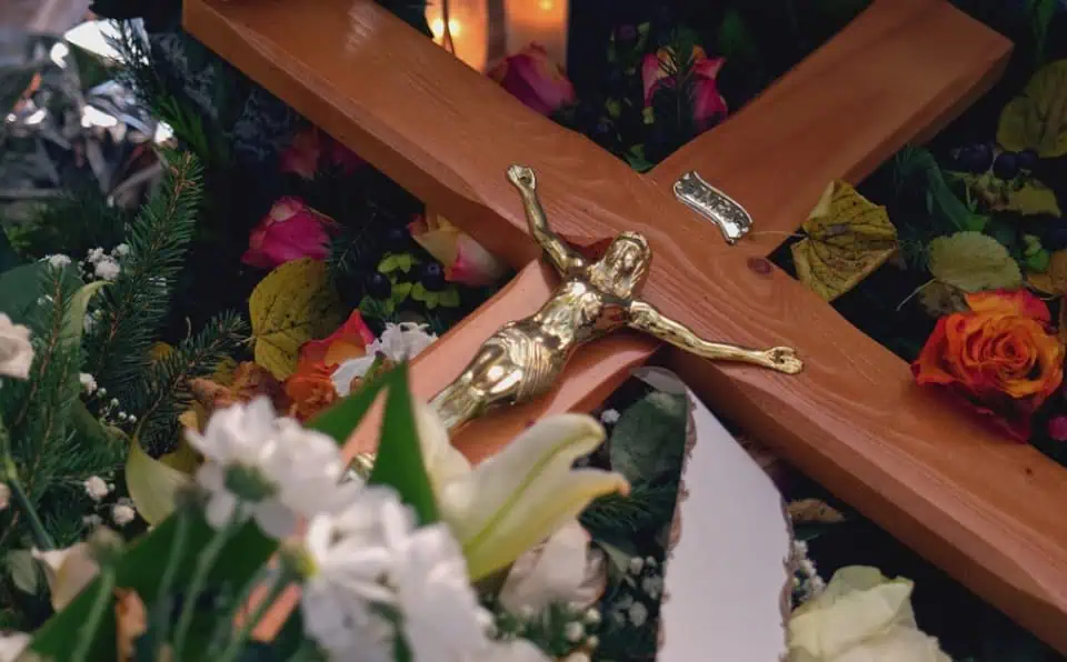 Close-up of a gold crucifix on a wooden cross with flowers around it.