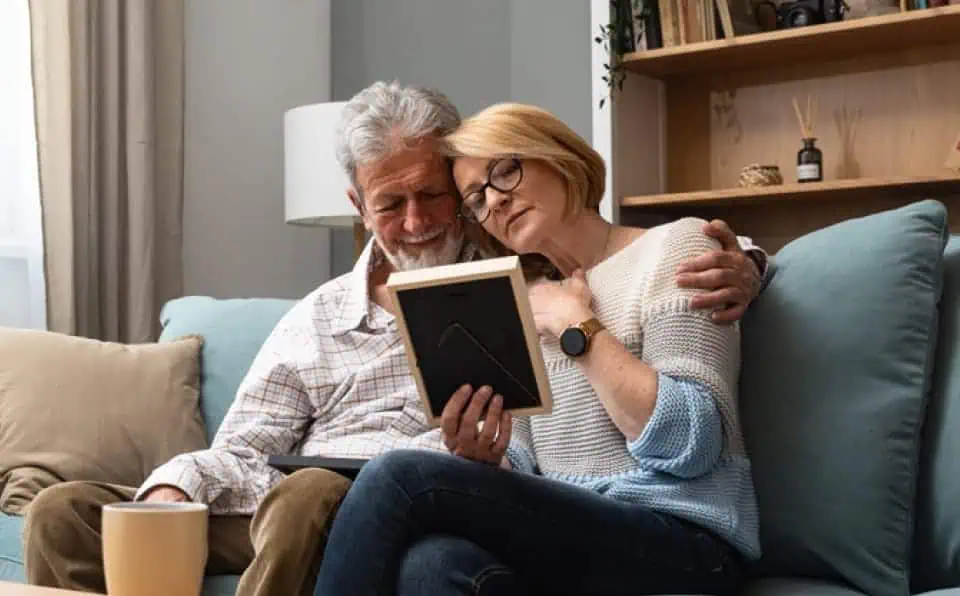 Elderly couple sitting on a sofa, looking at a framed portrait together.