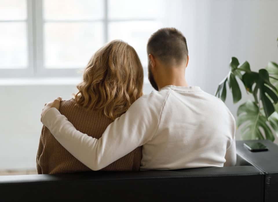 Two people sitting on a bench, embracing, in a bright room with large windows and a plant.