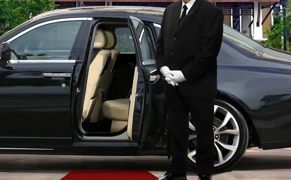 A chauffeur in formal attire standing next to a black limousine with open door.
