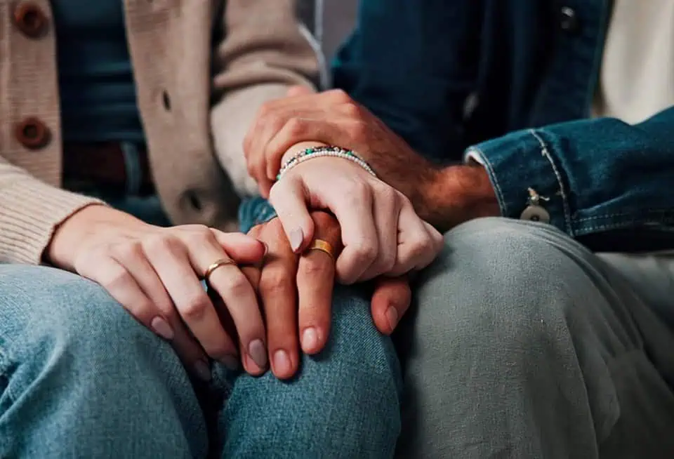 Two people holding hands, one with a ring and bracelet, the other with a ring, showing support.