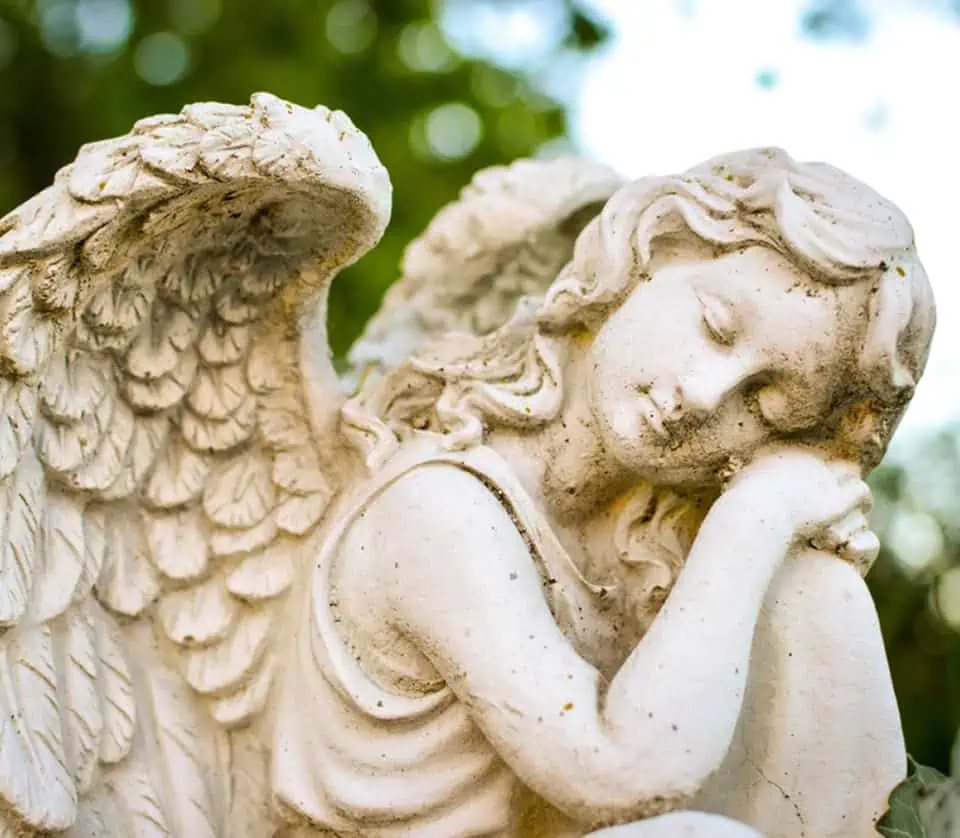 Stone angel statue with closed eyes and folded hands in a cemetery setting.