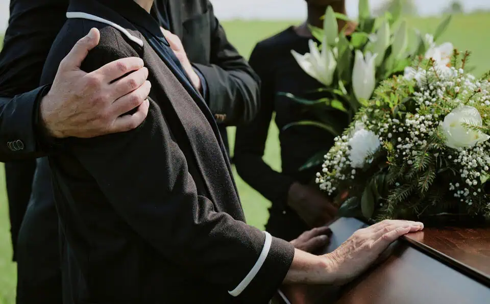 People gathered outdoors for a funeral service, with a focus on a person holding a hand on their own.