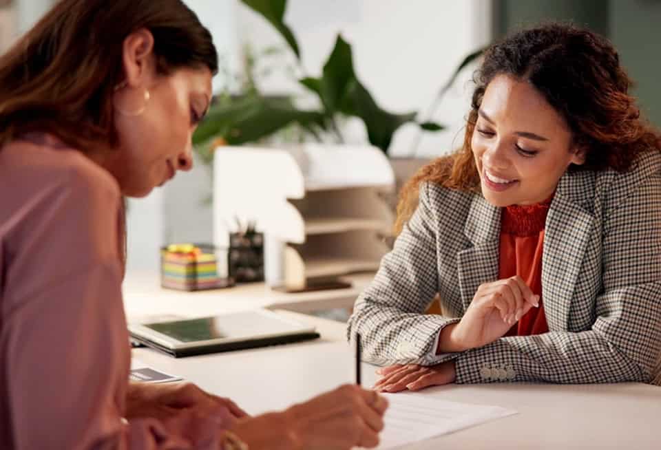 Two women discussing documents at a desk in an office setting.