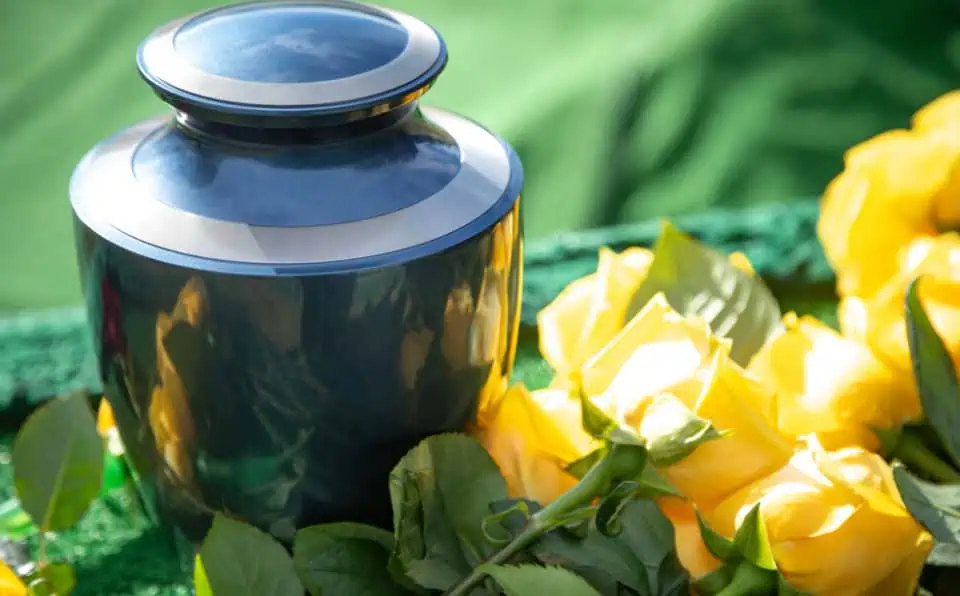 A polished black memorial urn surrounded by yellow roses on a green surface.