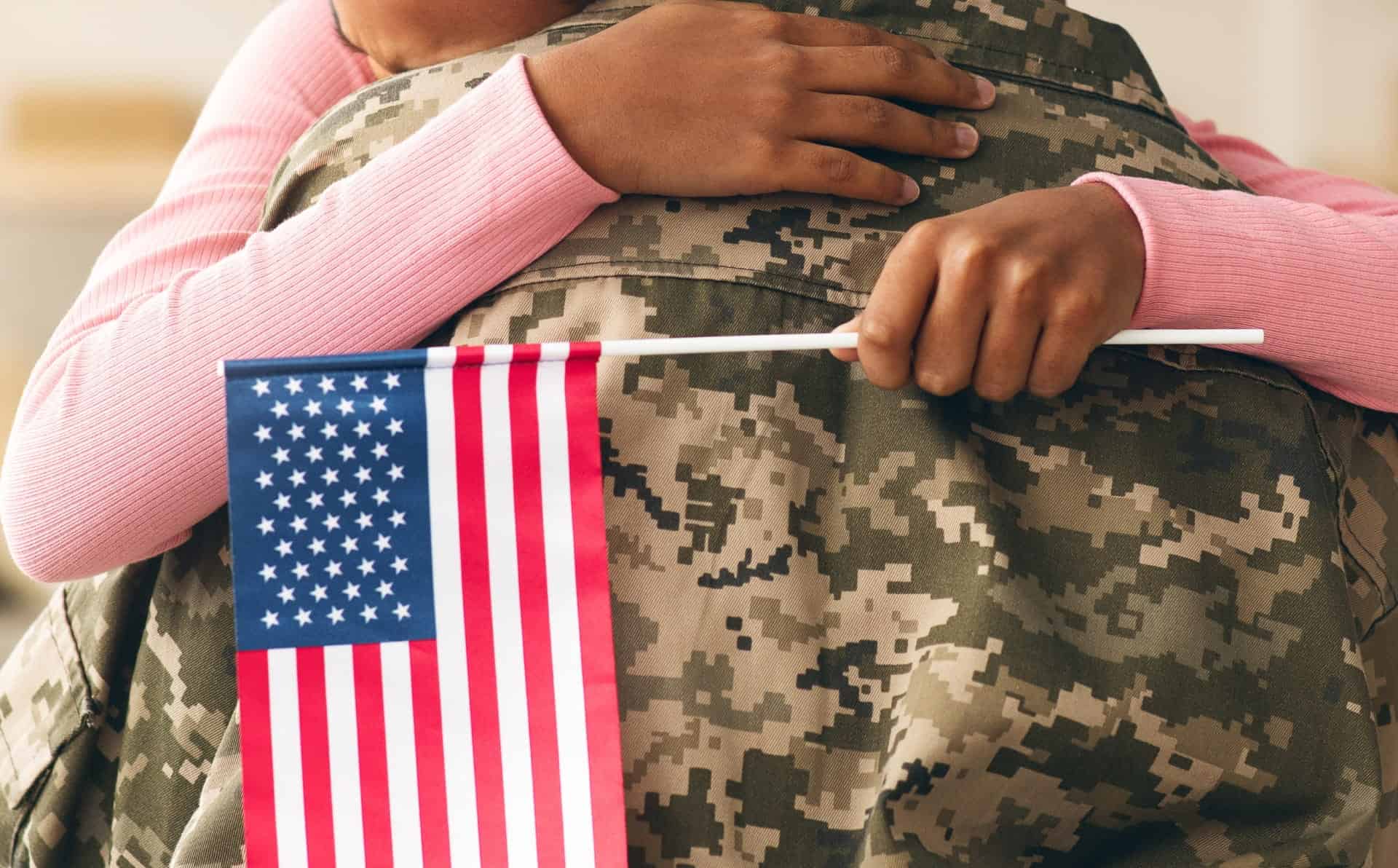 Person in pink sweater hugging a soldier in camouflage uniform with American flag.