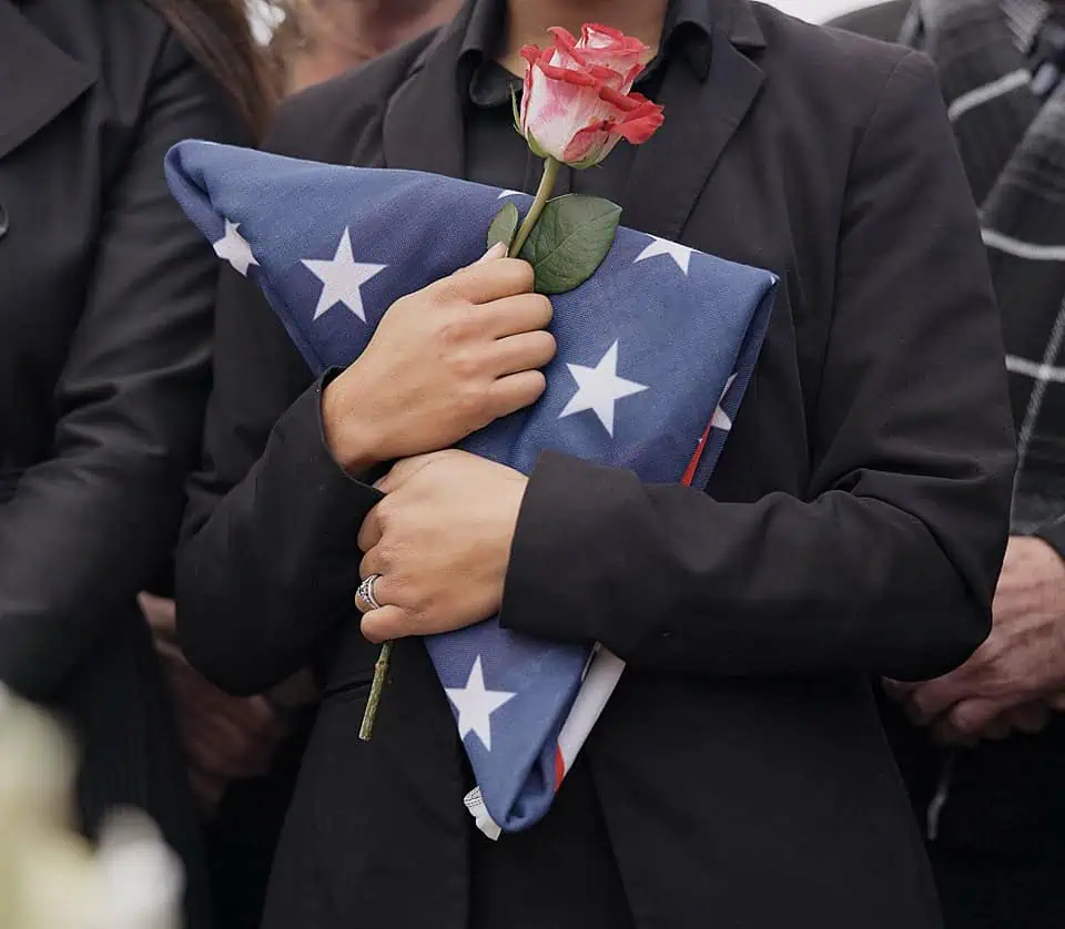 Person holding folded American flag and a pink rose at a memorial event.