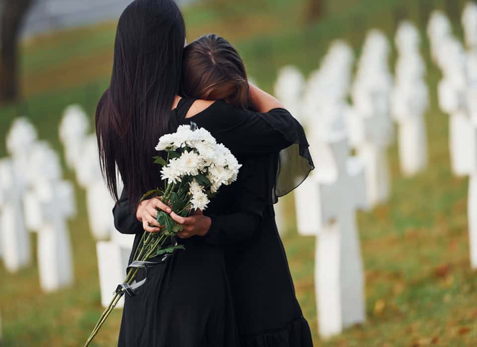 Two women hugging at a cemetery with white crosses in the background.