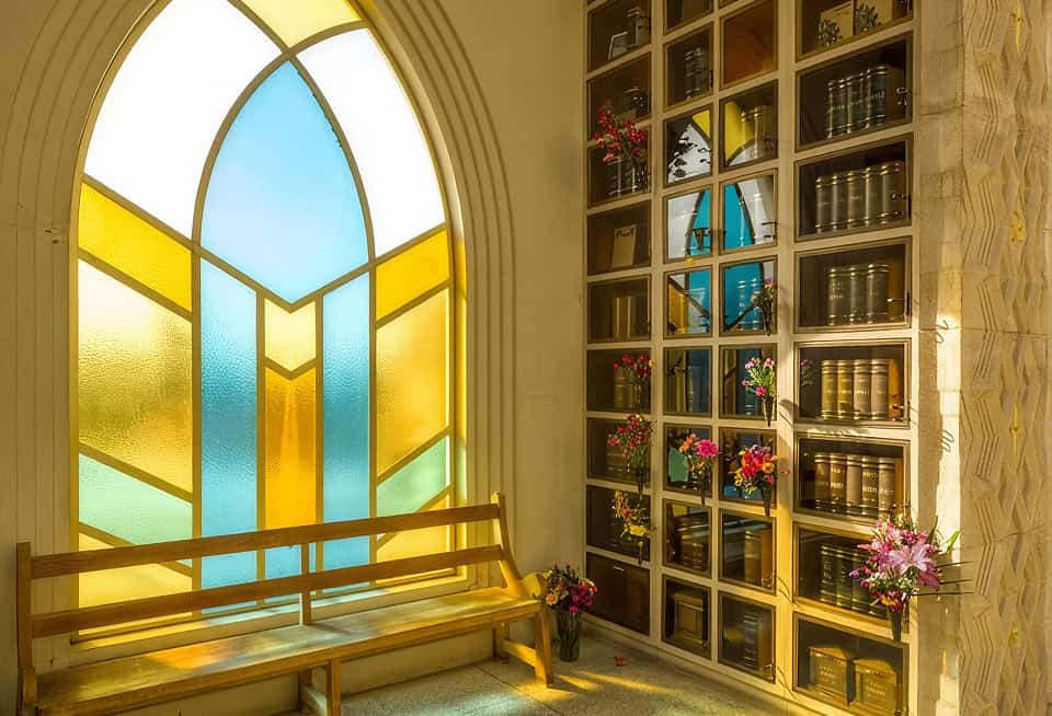 Bright stained glass window with a wooden bench and a tall bookshelf filled with books and flowers.