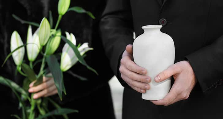 Person holding a white cremation urn with lilies in the background.
