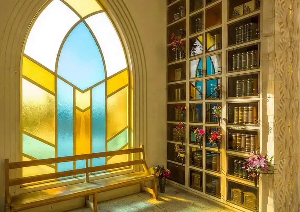 Sunlight streams through a stained glass window illuminating a wooden bench and a tall bookcase.