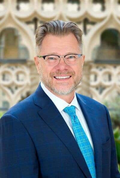 Man in a blue suit and glasses smiling outdoors with decorative architecture behind him.