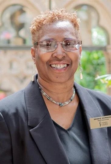 Portrait of a smiling woman with glasses and short curly hair, wearing a black blazer and name tag.