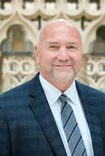 A middle-aged man with a beard and bald head, wearing a suit and tie, standing outdoors.