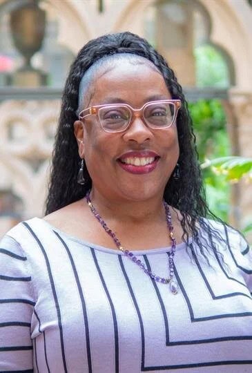 Smiling woman with glasses and jewelry in a garden setting.