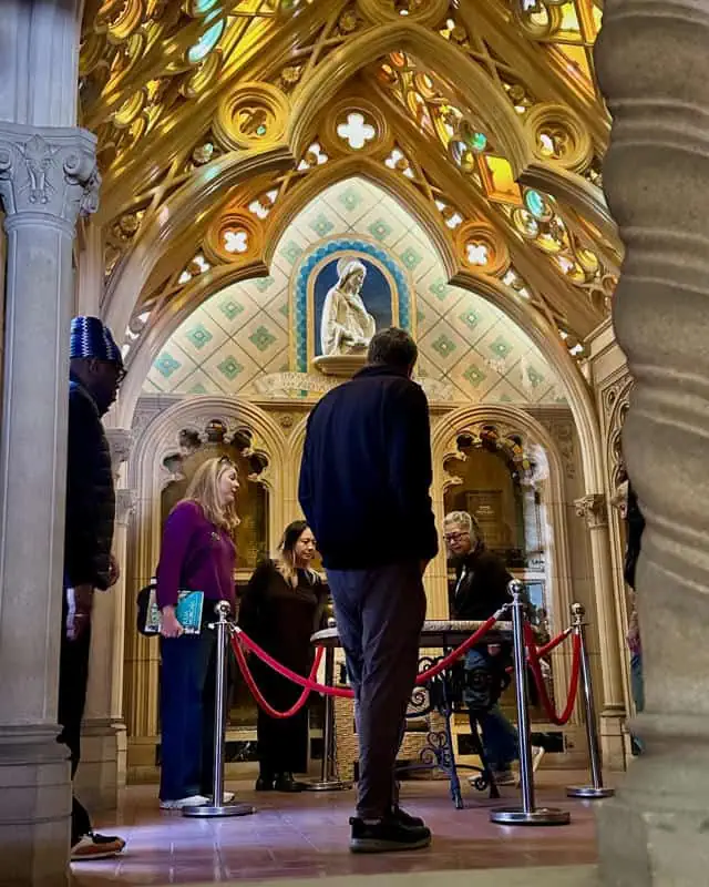 Chapel interior with visitors near red ropes and a statue in a stained glass arch.